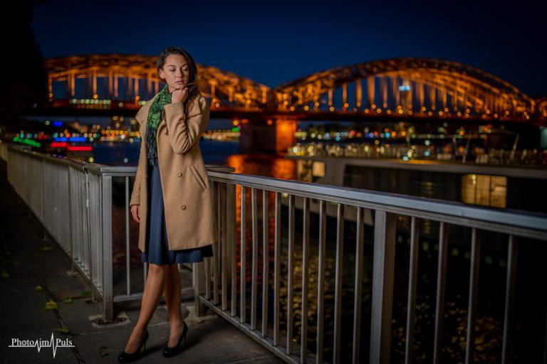 Frau im Mantel an einem Geländer, im Hintergrund eine beleuchtete Brücke bei Nacht