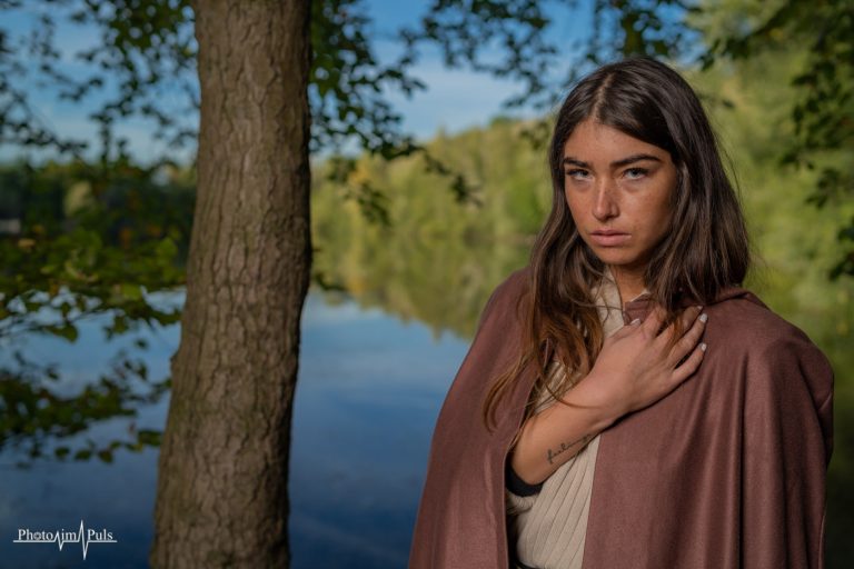 Junge Frau mit braunem Umhang an einem Waldsee