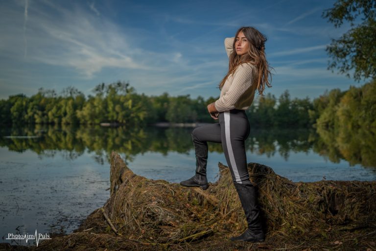 Frau in Jeans steht auf einem Baumstamm am See im Abendlicht