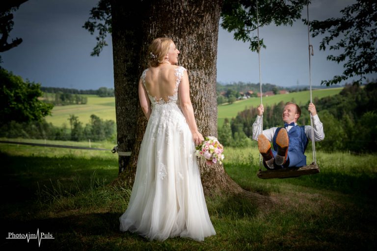 Braut lehnt an einem großen Baum, der Bräutigam sitzt daneben auf einer Schaukel