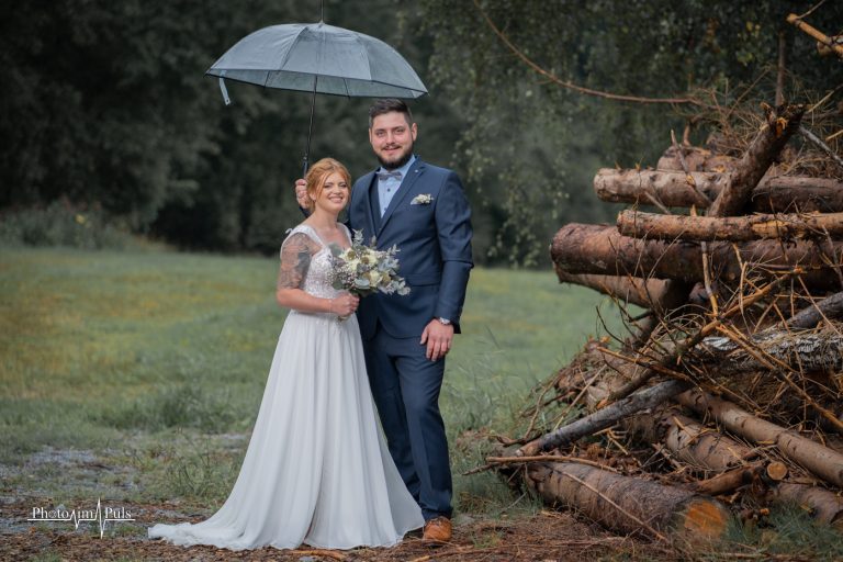 Brautpaar unter einem Regenschirm neben gestapeltem Holz