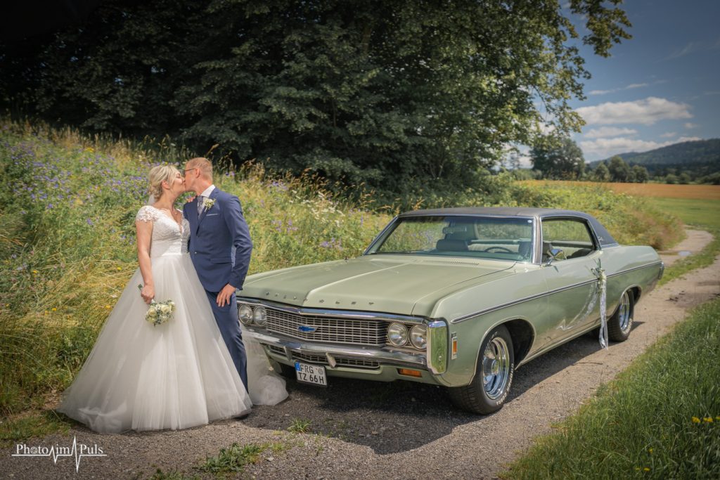 Brautpaar steht neben einem grünen Oldtimer auf einem Feldweg